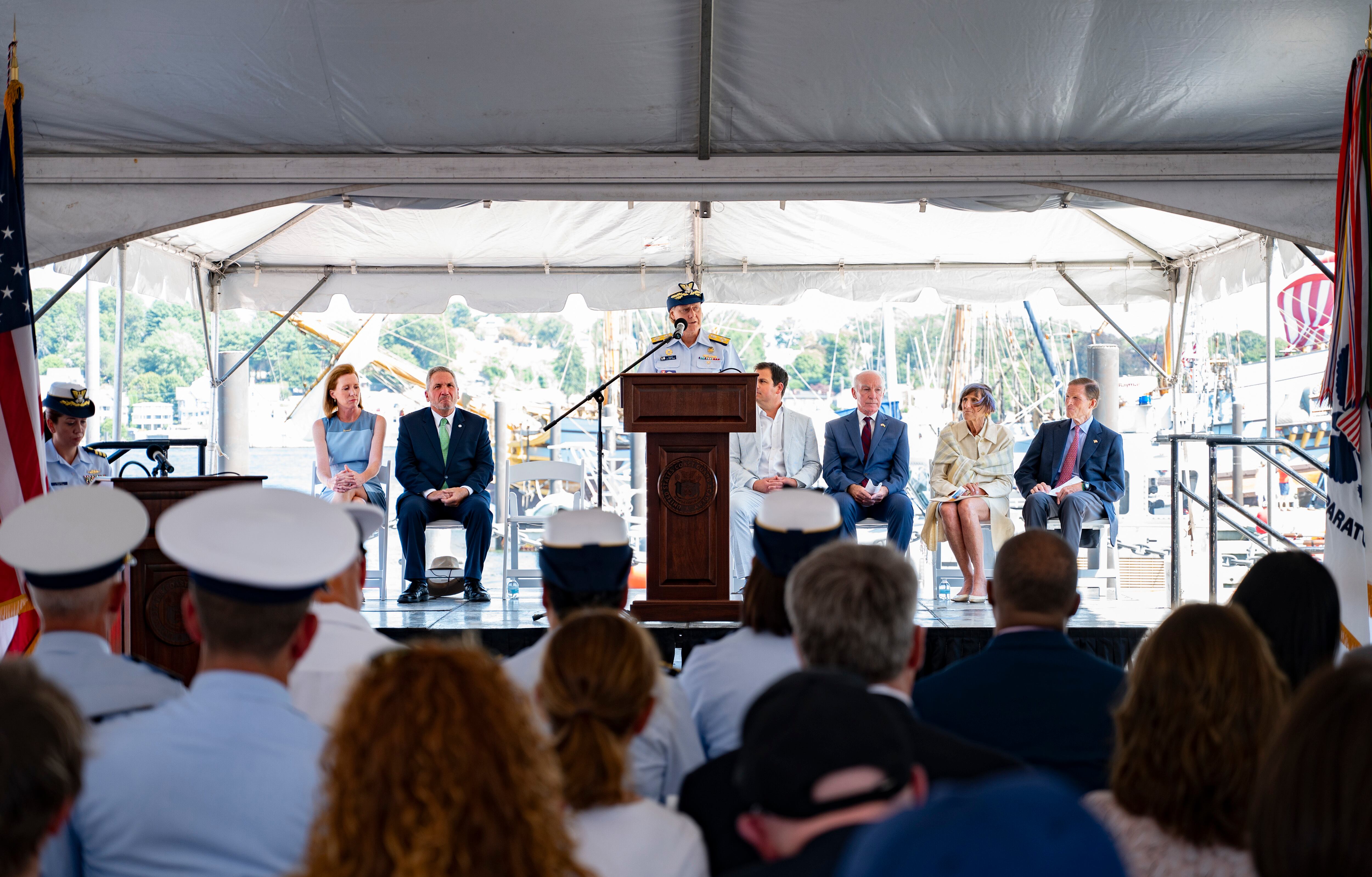 Adm. Linda Fagan speaks at a podium, flanked by seated lawmakers and museum officials, to an outdoor gathering of guests for the keel-laying ceremony.